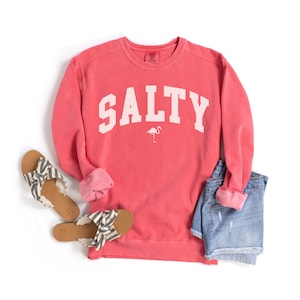 Op de afbeelding: Een koraalroze sweatshirt met het woord "SALTY" in wit op de voorkant. Een kleine roze flamingo-afbeelding bevindt zich onder het woord. Het sweatshirt ligt op een witte achtergrond met een paar zwart-witte sandalen en een spijkerbroek.