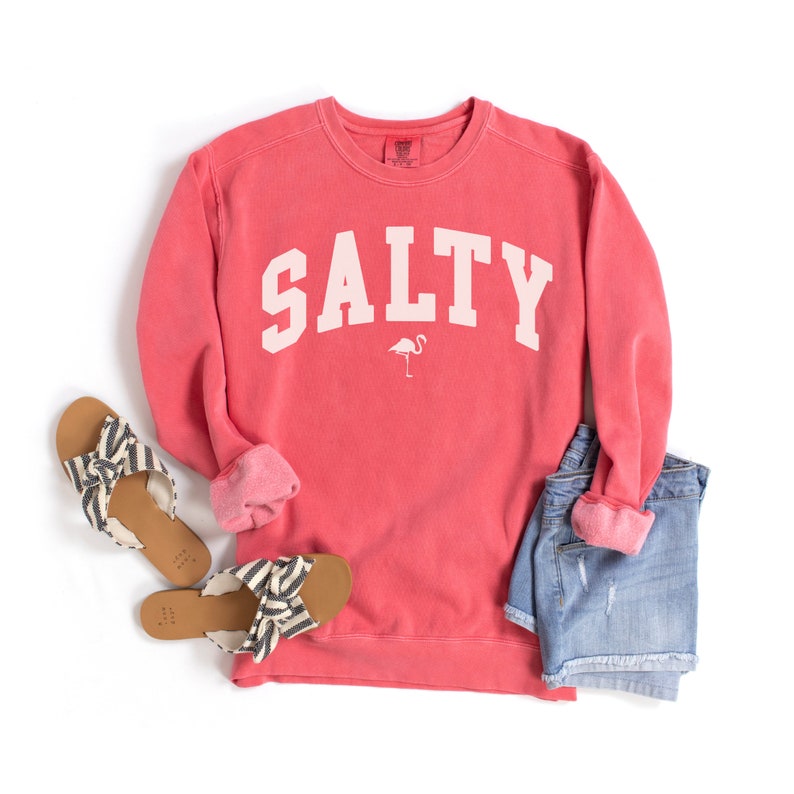 Op de afbeelding: Een koraalroze sweatshirt met het woord "SALTY" in wit op de voorkant. Een kleine roze flamingo-afbeelding bevindt zich onder het woord. Het sweatshirt ligt op een witte achtergrond met een paar zwart-witte sandalen en een spijkerbroek.