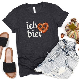 Op de afbeelding: Een donkergrijs T-shirt met de tekst "ich &hearts; bier" in witte letters en een bruine pretzel in de vorm van een hart.