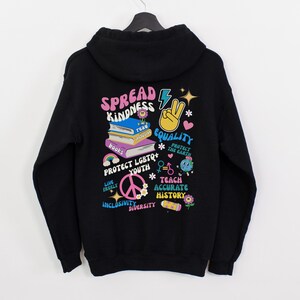 May include: Black hoodie with a colorful graphic print that says "Spread Kindness" and includes images of books, a peace sign, a rainbow, and a lightning bolt. The graphic also includes the words "Protect LGBTQ+ Youth", "Teach Accurate History", "Live Freely", "Inclusivity", "Diversity", and "Protect the Earth".
