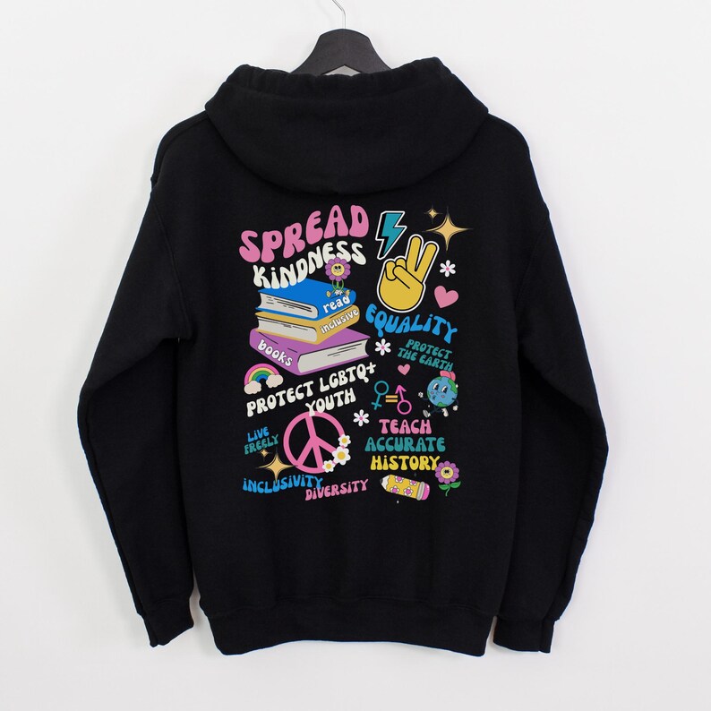May include: Black hoodie with a colorful graphic print that says "Spread Kindness" and includes images of books, a peace sign, a rainbow, and a lightning bolt. The graphic also includes the words "Protect LGBTQ+ Youth", "Teach Accurate History", "Live Freely", "Inclusivity", "Diversity", and "Protect the Earth".