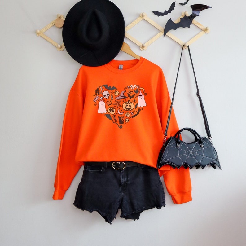 Op de afbeelding: Oranje sweatshirt met een hartvormig ontwerp met verschillende Halloween-iconen, waaronder geesten, pompoenen, vleermuizen en een heksenhoed. Het sweatshirt wordt gecombineerd met een zwarte denim short en een zwarte vleermuisvormige handtas.