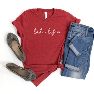 K&ouml;nnte beinhalten: Rotes T-Shirt mit dem Text "lake life" in Wei&szlig; mit einem kleinen wei&szlig;en Herz. Das Shirt liegt auf einem wei&szlig;en Hintergrund mit einem Paar grauen Ballerinas und einer blauen Jeans.