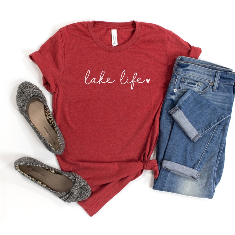 K&ouml;nnte beinhalten: Rotes T-Shirt mit dem Text "lake life" in Wei&szlig; mit einem kleinen wei&szlig;en Herz. Das Shirt liegt auf einem wei&szlig;en Hintergrund mit einem Paar grauen Ballerinas und einer blauen Jeans.
