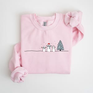 Op de afbeelding: Een lichtroze sweatshirt met een ronde hals. De voorkant heeft een grillige illustratie van drie sneeuwpoppen en een kerstboom in zwart en wit. De middelste sneeuwpop draagt een rode hoed en sjaal. Sneeuwvlokken vallen.