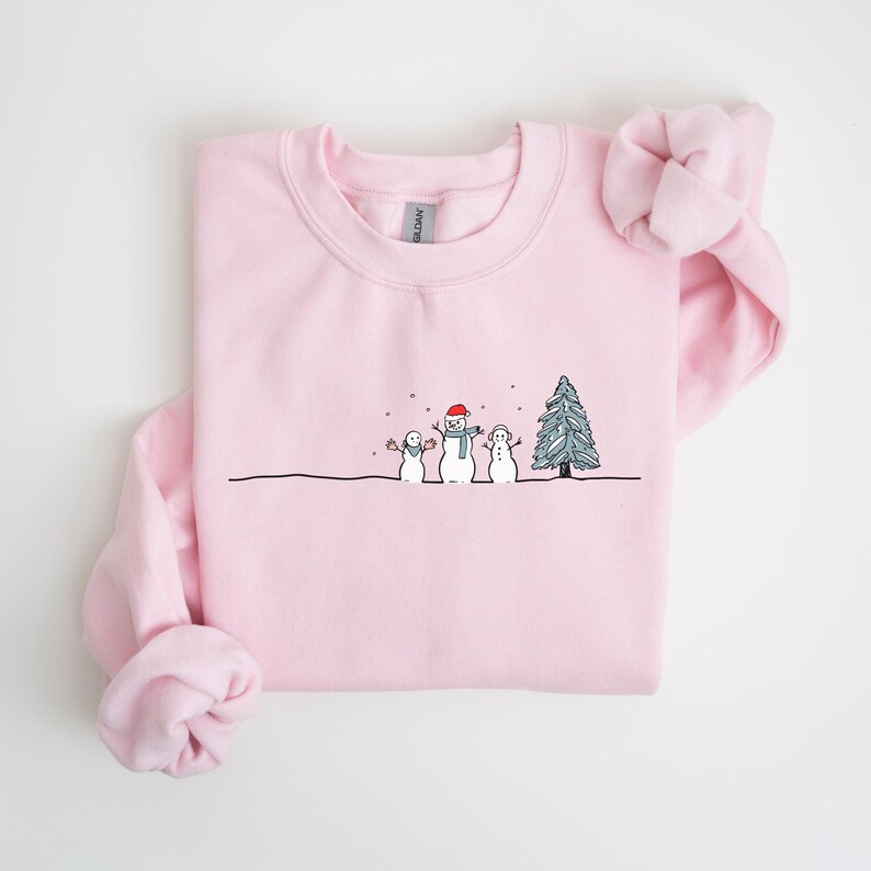 Op de afbeelding: Een lichtroze sweatshirt met een ronde hals. De voorkant heeft een grillige illustratie van drie sneeuwpoppen en een kerstboom in zwart en wit. De middelste sneeuwpop draagt een rode hoed en sjaal. Sneeuwvlokken vallen.