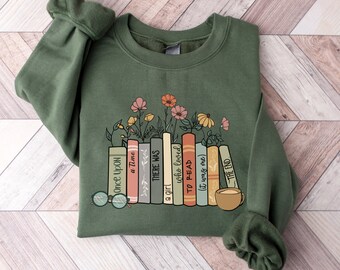 Può includere: Una felpa verde con una grafica di una pila di libri con fiori che crescono da essi. I libri hanno titoli che recitano "Once Upon a Time", "There Was", "a girl who loved", "To Read", "(it was me)" e "The End".