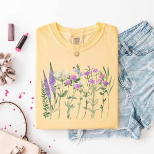 May include: Yellow Comfort Colors t-shirt with a watercolor floral design featuring purple, lavender, and white wildflowers. The shirt is paired with denim shorts, a necklace, and makeup.