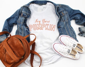 May include: A white t-shirt with the text "Hey there PUMPKIN" in orange, paired with a denim jacket, brown leather backpack, and white Converse sneakers. The outfit is laid out on a white surface.