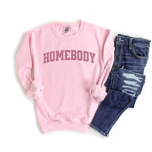 Puede incluir: Sudadera rosa con la palabra "HOMEBODY" impresa en color granate en una fuente de estilo universitario.