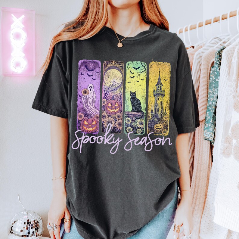 May include: Dark gray t-shirt with a colorful Halloween-themed graphic. The design features a ghost, pumpkins, a black cat, and a spooky castle, with the words "Spooky Season" in a cursive font.