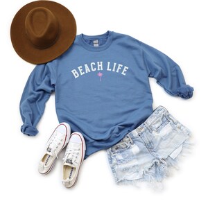 Pode incluir: Uma camisa de frio azul com o texto "Beach Life" em branco e um gr&aacute;fico de palmeira rosa. A camisa de frio &eacute; combinada com t&ecirc;nis brancos, shorts jeans e um chap&eacute;u fedora marrom.