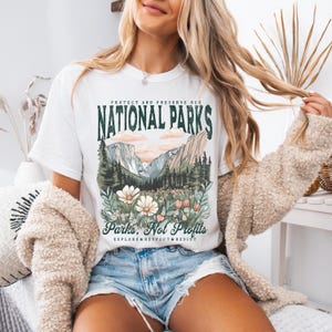 K&ouml;nnte beinhalten: Wei&szlig;es T-Shirt mit einem grafischen Aufdruck einer Berglandschaft und Blumen. Der Text "Protect and Preserve Our National Parks" ist in Gr&uuml;n &uuml;ber dem Bild gedruckt. Unter dem Bild ist der Text "Parks, Not Profits" in Gr&uuml;n gedruckt. Der Text "Explore. Respect. Resist" ist in kleineren gr&uuml;nen Buchstaben unter dem Haupttext gedruckt.
