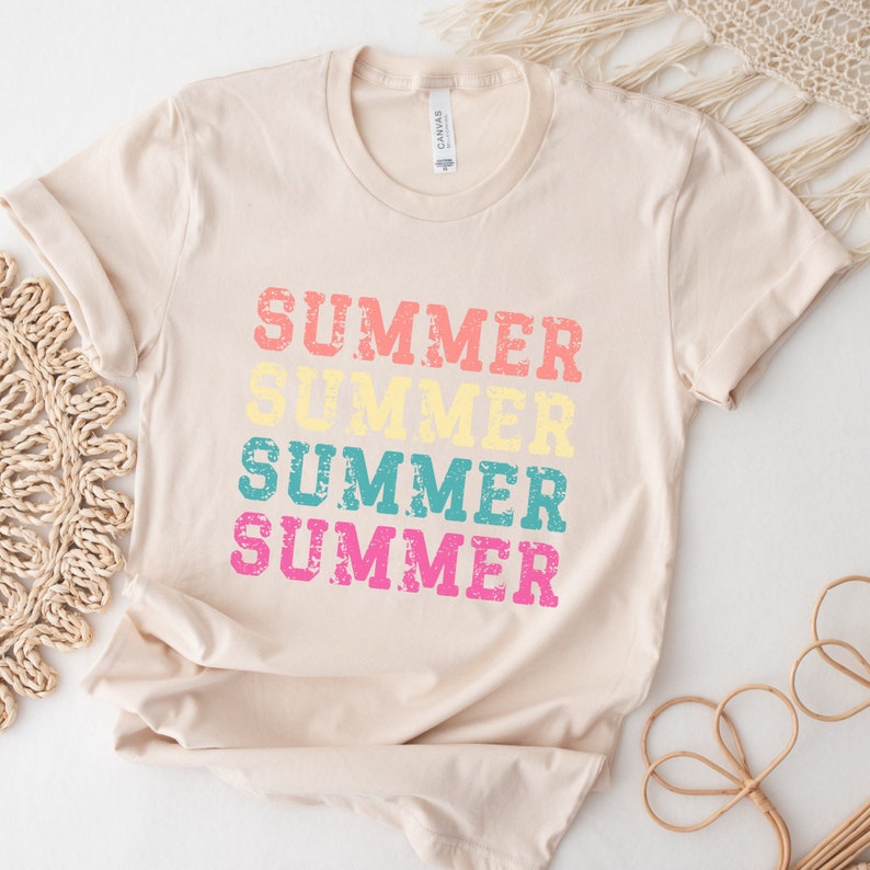 May include: A cream-colored t-shirt with the word "SUMMER" printed four times in a stacked design. The word is in a distressed font, with each instance in a different color: coral, yellow, teal, and pink. The shirt has short sleeves and a crew neck.