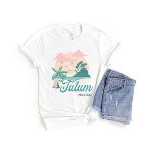 May include: White t-shirt with a graphic of a palm tree, surfboard, and sunset with the text "Tulum Mexico".
