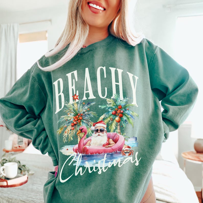 May include: Green sweatshirt with the words "BEACHY Christmas" in white. The graphic features Santa Claus in sunglasses and a Santa hat, floating in a pink flamingo pool, with palm trees and coconuts.