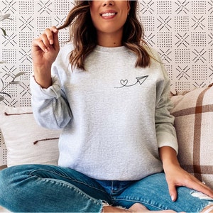 Op de afbeelding: Een grijs sweatshirt met een kleine zwarte afbeelding van een papieren vliegtuigje met een hartje aan de staart. Het sweatshirt wordt gedragen door iemand die op een bank zit.