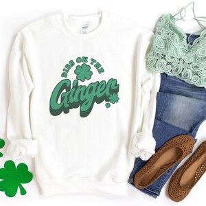K&ouml;nnte beinhalten: Ein wei&szlig;es Sweatshirt mit dem gr&uuml;nen Schriftzug "Dibs on the Ginger" und Kleeblatt-Akzenten. Das Bild zeigt auch braune Ballerinas, eine blaue Jeans und ein gr&uuml;nes Spitzentop. Das Sweatshirt hat ein St. Patrick's Day-Thema.