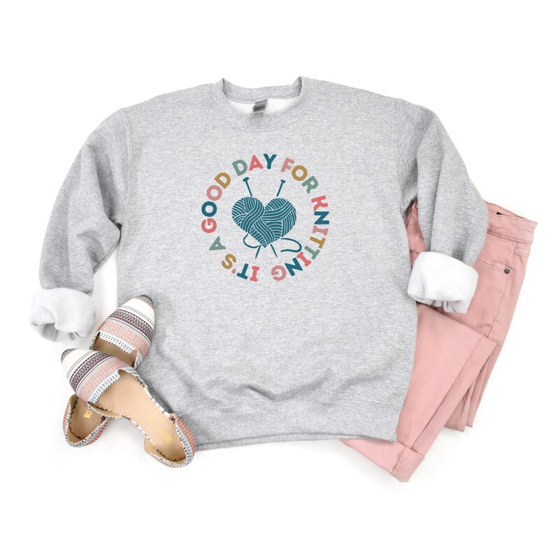 K&ouml;nnte beinhalten: Ein graues Sweatshirt mit dem Text "Es ist ein guter Tag zum Stricken" in einem Kreis um eine herzf&ouml;rmige Wollkn&auml;uel mit Stricknadeln.