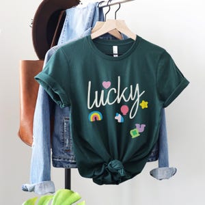 Puede incluir: Una camiseta verde oscuro con la palabra "lucky" escrita en blanco. La camiseta tiene un arco&iacute;ris, un unicornio, una estrella, un globo aerost&aacute;tico y un tr&eacute;bol de cuatro hojas.