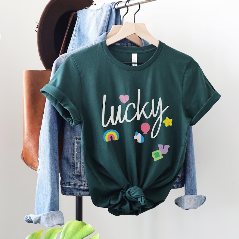 Puede incluir: Una camiseta verde oscuro con la palabra "lucky" escrita en blanco. La camiseta tiene un arco&iacute;ris, un unicornio, una estrella, un globo aerost&aacute;tico y un tr&eacute;bol de cuatro hojas.