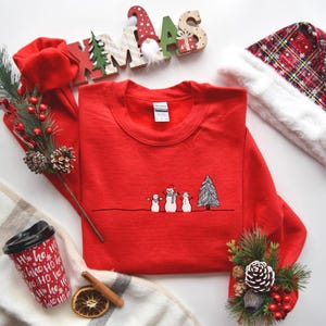 Op de afbeelding: Een rode crewneck sweatshirt met een lijntekening van drie sneeuwpoppen en een kerstboom. De sweatshirt is omgeven door kerstversieringen, waaronder een kerstmuts, bessen en een koffiekopje.