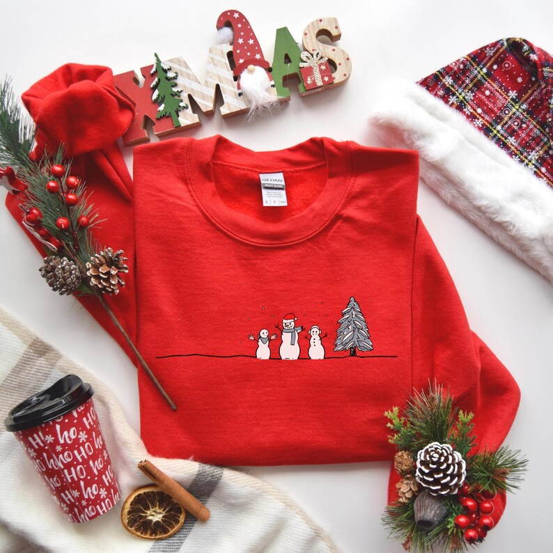 Op de afbeelding: Een rode crewneck sweatshirt met een lijntekening van drie sneeuwpoppen en een kerstboom. De sweatshirt is omgeven door kerstversieringen, waaronder een kerstmuts, bessen en een koffiekopje.