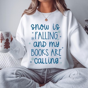 May include: White sweatshirt with the text "snow is falling and my books are calling" in blue script. The design includes snowflakes. A person is holding a mug with a Christmas tree design.