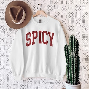 Może przedstawiać: Biała bluza z czerwonym nadrukiem "SPICY" z przodu. Bluza wisi na wieszaku na białym tle z czarno-białym wzorem.