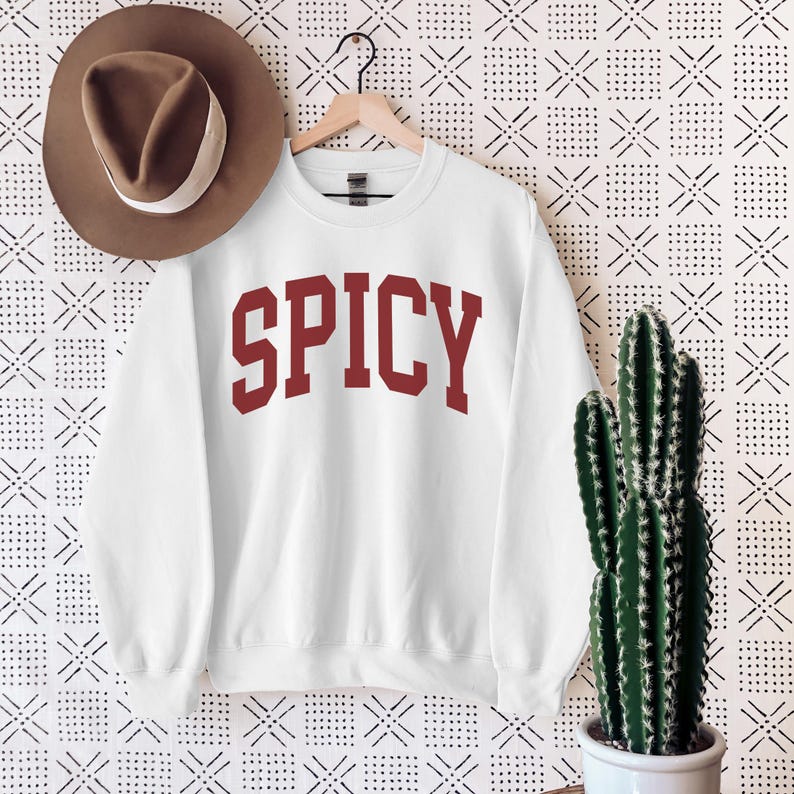 Może przedstawiać: Biała bluza z czerwonym nadrukiem "SPICY" z przodu. Bluza wisi na wieszaku na białym tle z czarno-białym wzorem.