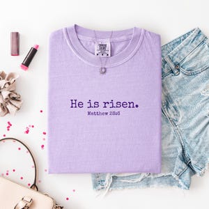 May include: A lilac Comfort Colors t-shirt with the text "He is risen." and Matthew 28:6 printed in purple. The shirt is folded and displayed with denim shorts, a necklace, makeup, and a scrunchie.