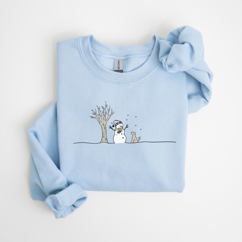 Op de afbeelding: Lichtblauw sweatshirt met een grillige illustratie van een sneeuwman, een hond en een kale boom. De sneeuwman draagt een hoed en sjaal. De hond zit naast de sneeuwman. Sneeuwvlokken vallen. Het sweatshirt is gemaakt van een zacht materiaal.