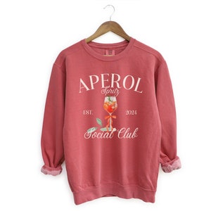 Può includere: Una felpa rossa con un design grafico bianco che dice "Aperol Spritz Social Club" con un'illustrazione di un bicchiere di Aperol Spritz. Il testo dice anche "Est. 2024".