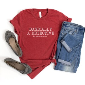 May include: Red t-shirt with the text "BASICALLY A DETECTIVE #truecrimejunkie" in white. The shirt is laid out on a white surface with a pair of grey flats and blue jeans.