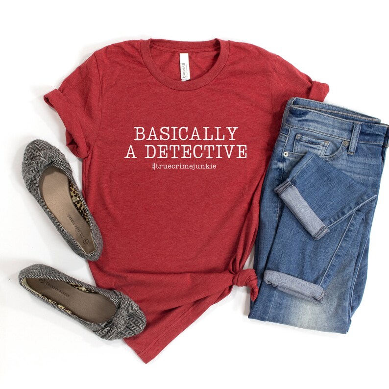 May include: Red t-shirt with the text "BASICALLY A DETECTIVE #truecrimejunkie" in white. The shirt is laid out on a white surface with a pair of grey flats and blue jeans.