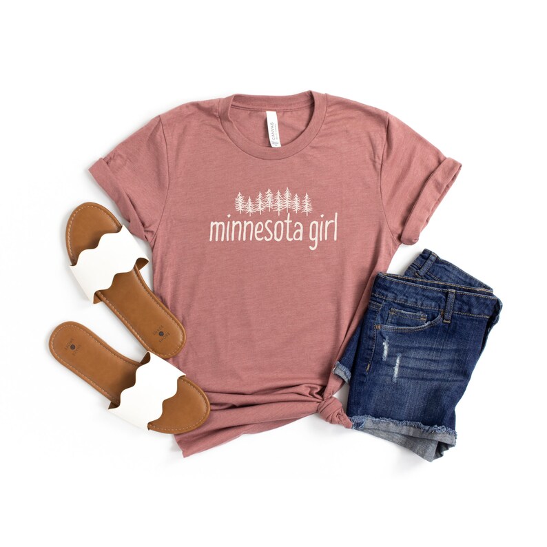 May include: A mauve t-shirt with a white graphic of a line of pine trees and the text "minnesota girl". The t-shirt is laid out on a white background with a pair of white sandals and denim shorts.