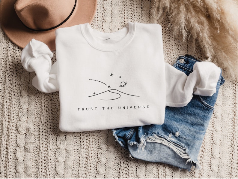 Op de afbeelding: Wit sweatshirt met een zwarte lijntekening van een planeet, sterren en een bergketen. De tekst "TRUST THE UNIVERSE" is onder de tekening gedrukt.
