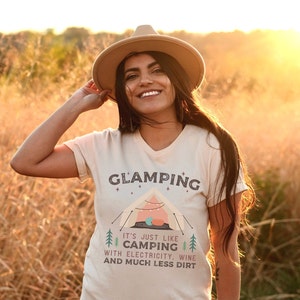 May include: A woman wearing a beige t-shirt with the text "Glamping It's just like camping with electricity, wine and much less dirt" and a brown hat. The shirt features a cartoon illustration of a tent with fairy lights.