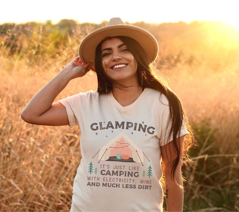 May include: A woman wearing a beige t-shirt with the text "Glamping It's just like camping with electricity, wine and much less dirt" and a brown hat. The shirt features a cartoon illustration of a tent with fairy lights.