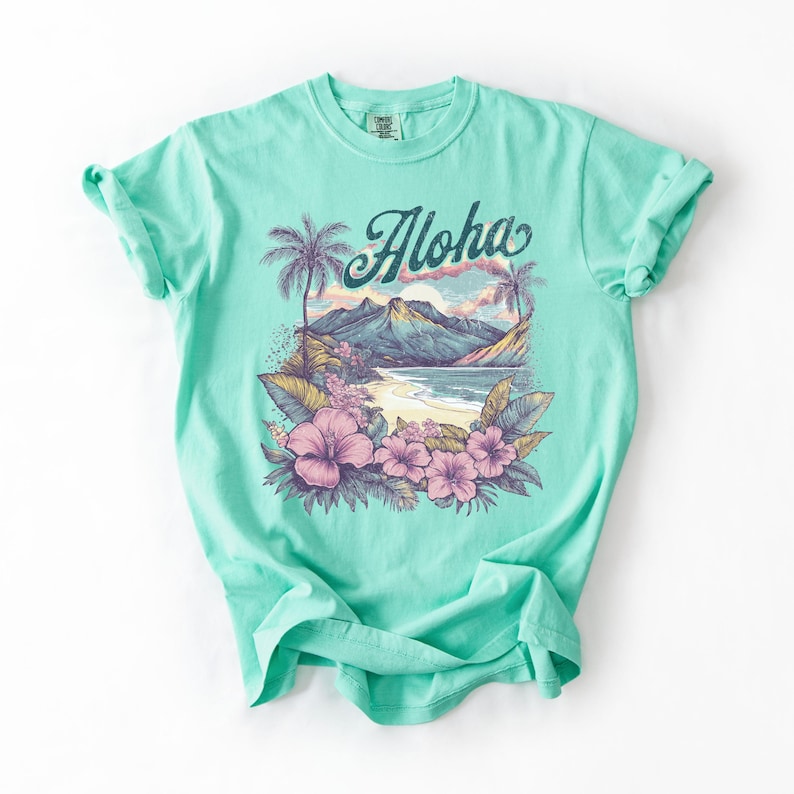 以下が含まれることがあります： トロピカルな風景デザインのターコイズ色のTシャツ。グラフィックには、山、ヤシの木、花のあるビーチシーンが含まれており、「Aloha」の文字が書かれています。シャツは柔らかい素材で作られています。