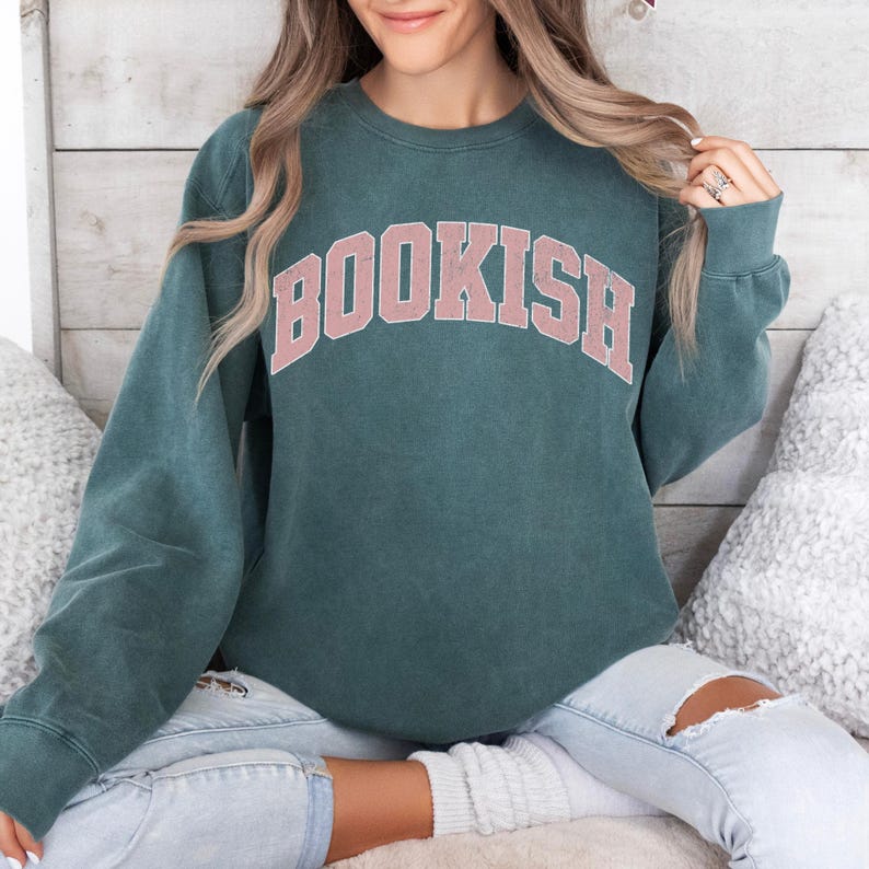 Puede incluir: Una sudadera azul verdosa con un gr&aacute;fico rosa "BOOKISH" en una fuente universitaria.