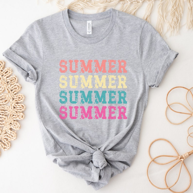 May include: A gray t-shirt with the word "SUMMER" printed four times in a stacked design. The word is in a distressed font and features the colors peach, yellow, teal, and pink. The shirt is knotted at the waist.