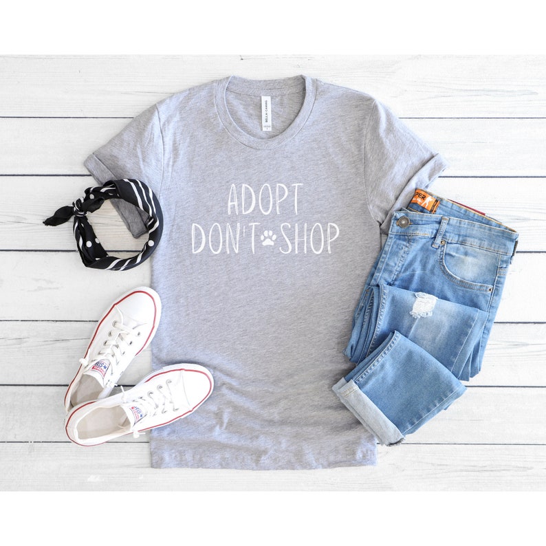 May include: A gray t-shirt with the text "Adopt Don't Shop" in white with a paw print symbol. The shirt is laid out on a white wooden surface with a pair of white sneakers, a black and white bandana, and a pair of blue jeans.