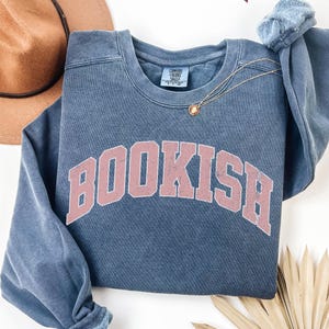 Puede incluir: Una sudadera azul con la palabra "BOOKISH" impresa en rosa en la parte delantera. La sudadera tiene un aspecto desgastado y est&aacute; hecha de una tela suave y c&oacute;moda.