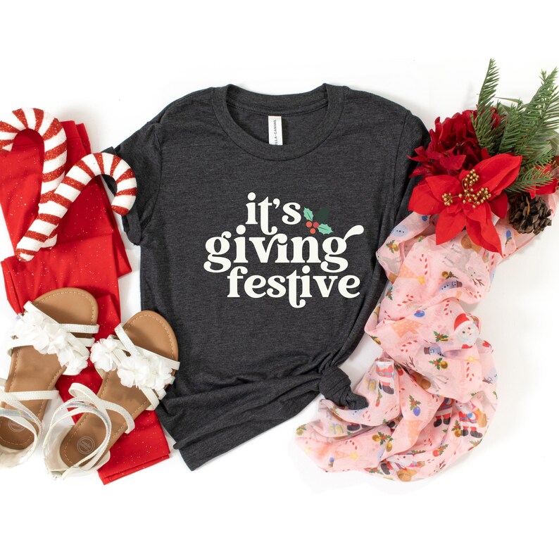 May include: A dark gray t-shirt with the text "it's giving festive" in white. The image includes red leggings, white sandals, candy canes, and a pink scarf with a holiday print. A bouquet of red flowers and pinecones is also present.