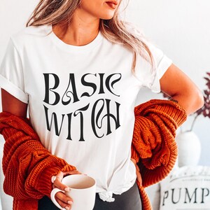 May include: A white t-shirt with the text "BASIC WITCH" printed in black. The shirt is being worn by a person with long blonde hair and a brown cardigan.