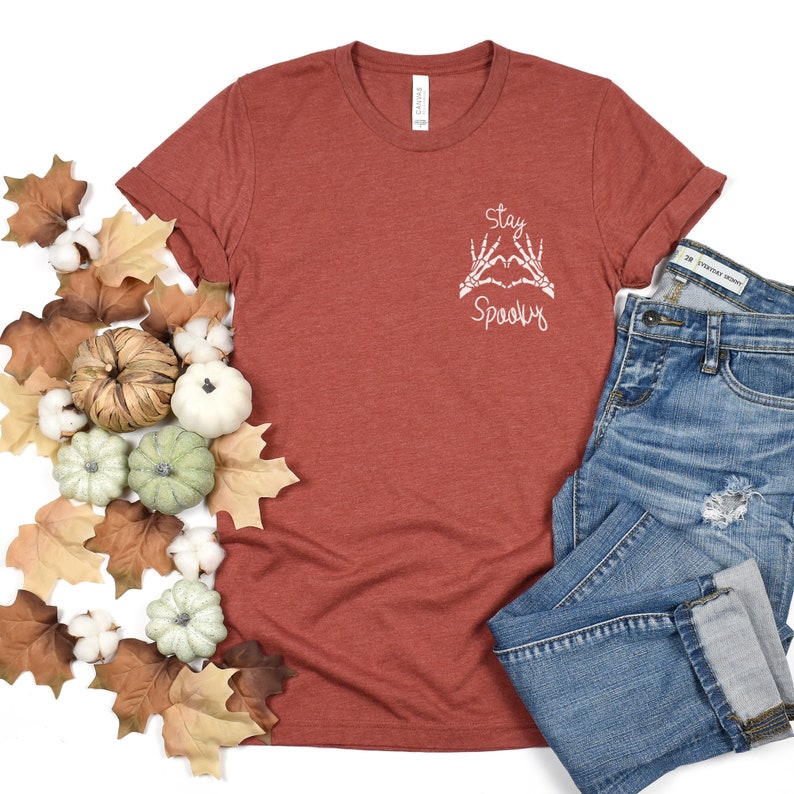 May include: A rust-colored t-shirt with a white graphic of skeleton hands making a heart shape with the text "Stay Spooky". The shirt is laying on a white surface with fall leaves and cotton balls around it. A pair of blue jeans is also in the image.