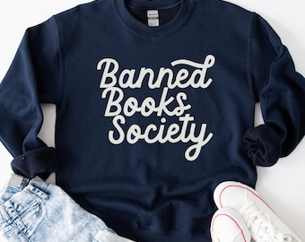 May include: Navy blue sweatshirt with the text "Banned Books Society" in white script. The sweatshirt is paired with light wash denim jeans and white canvas sneakers with red accents. The overall aesthetic is casual and book-themed.
