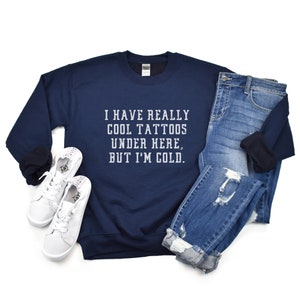 May include: A navy blue sweatshirt with the text "I have really cool tattoos under here, but I'm cold." printed in white on the front. The sweatshirt is paired with blue jeans and white sneakers.