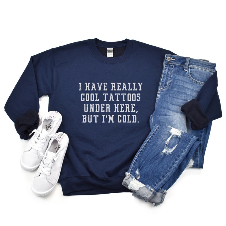 May include: A navy blue sweatshirt with the text "I have really cool tattoos under here, but I'm cold." printed in white on the front. The sweatshirt is paired with blue jeans and white sneakers.